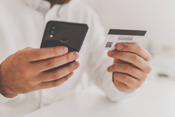 Die Hände des Mannes halten eine Kreditkarte, mit der er online einkaufen kann. Online-Bezahlung, Nahaufnahme eines jungen Mannes, der am Arbeitsplatz mit seinem Smartphone online einkauft. Web-Shopping . - Foto, Bild