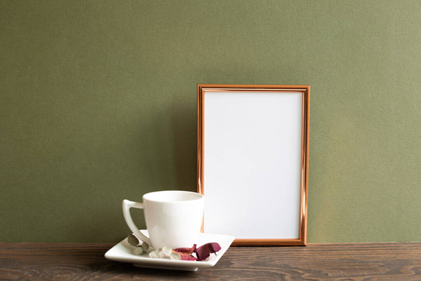 Пустая фоторамка с чашкой кофе и сухими цветами на деревянном столе на фоне хаки - Фото, изображение
