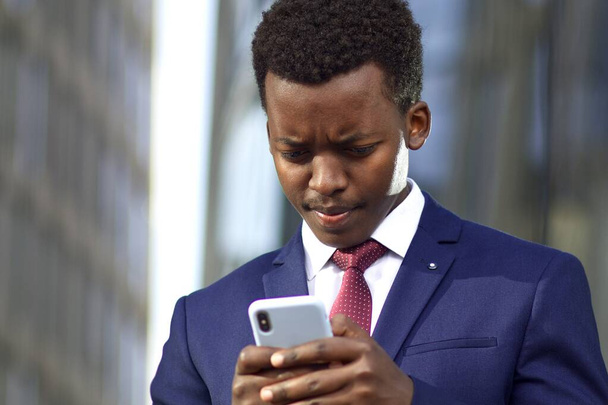 Serious young businessman in formal suit looking at screen of his cell mobile phone. Black African Afro American man typing a message on smartphone outdoors, frowning.  - Фото, зображення