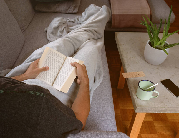Hombre en casa acogedora envuelto en blanco relajante en el sofá celebración de libro y lectura. Luz del sol caliente que entra en la sala de estar - Foto, imagen