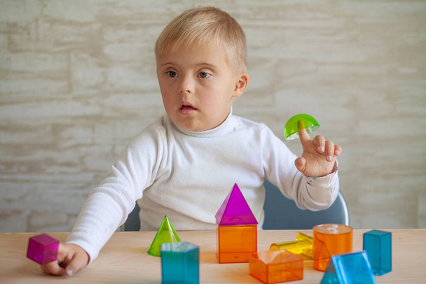 A genetikai betegséggel küzdő cuki kisfiúk ülnek az asztalnál. Down-szindrómás gyermek 3D-s geometriai alakokkal játszik. Műanyag játékokban. - Fotó, kép