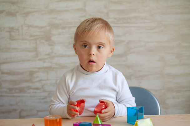 Pequeños chicos lindos con una enfermedad genética están sentados en la mesa. niño con síndrome de Down juega con figuras geométricas 3D. Plástico en juguetes. - Foto, Imagen