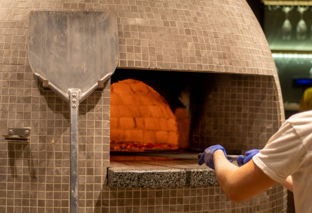 Руки изготовителя пиццы, который кладет пиццу в духовку лопатой. Огонь горит - Фото, изображение