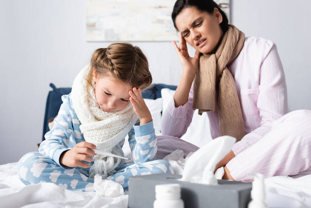 madre enferma e hija que sufren de dolor de cabeza mientras que la niña mirando el termómetro en primer plano borrosa - Foto, Imagen