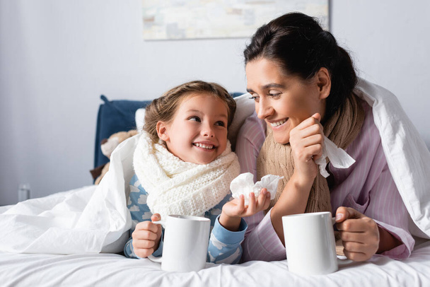 sick, cheerful mother and daughter looking at each other while holding cups with hot drink in bed - Fotoğraf, Görsel