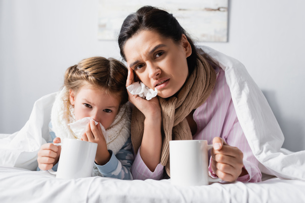 upset mother with child looking at camera while holding warm drink in bed - Foto, immagini
