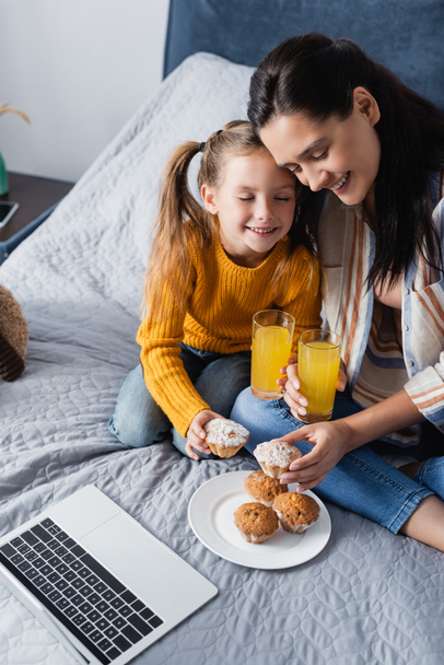 gelukkig moeder en dochter houden muffin en sinaasappelsap in de buurt van laptop - Foto, afbeelding