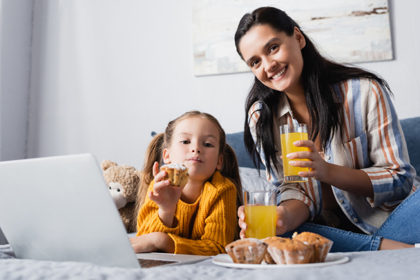 happy woman and daughter looking at camera near muffins and orange juice while watching film on laptop - Foto, immagini