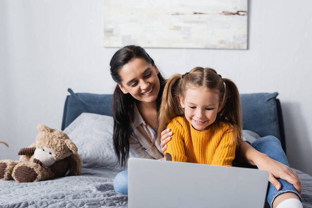 cheerful woman looking at daughter using laptop in bedroom - Fotó, kép
