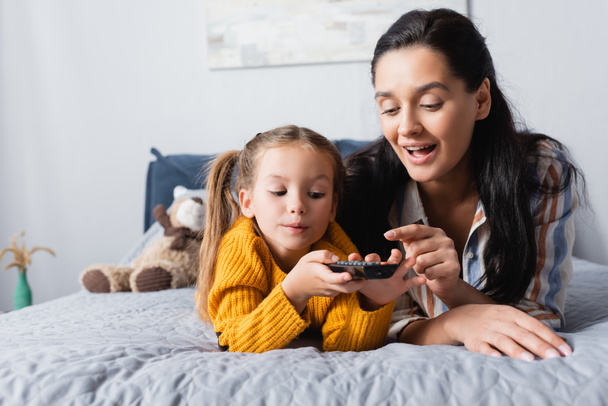 happy mother showing daughter how to use tv remote controller while lying on bed - Foto, Bild