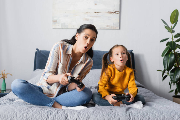 KYIV, UKRAINE - SEPTEMBER 15, 2020: shocked woman with daughter sitting with crossed legs and playing video game  - 写真・画像
