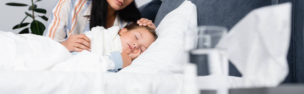 Mutter berührt kranke Tochter schlafend im Bett neben Wasserglas auf verschwommenem Vordergrund, Banner - Foto, Bild