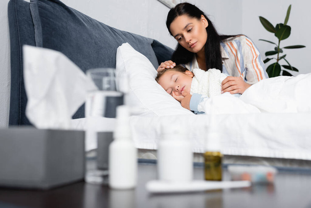 anya ölelés beteg lánya alszik az ágyban mellett éjjeliszekrény gyógyszerekkel elmosódott előtérben - Fotó, kép