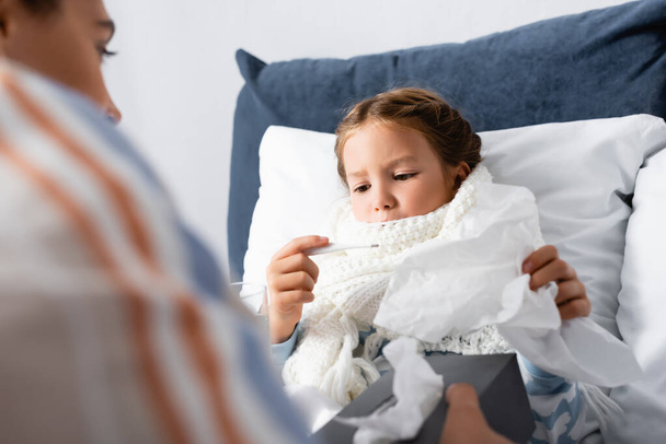 fille malade regardant thermomètre près de mère tenant des serviettes en papier sur le premier plan flou - Photo, image