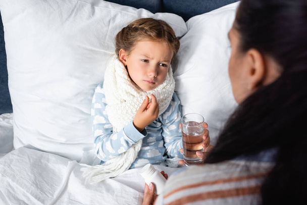 sick girl holding pill near mother holding glass of water on blurred foreground - Photo, Image