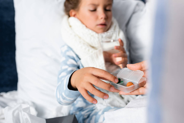 woman giving medicines to daughter lying in bed and holding glass of water on blurred background - Foto, Bild
