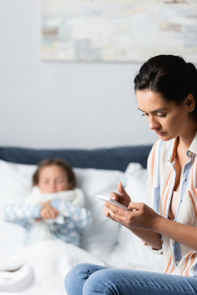 moeder messaging op mobiele telefoon in de buurt van zieke dochter liggend in bed op wazig achtergrond - Foto, afbeelding