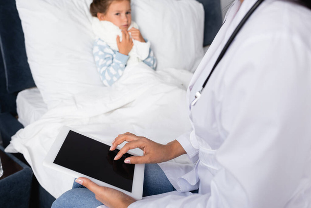 pédiatre à l'aide d'une tablette numérique avec écran vierge près d'une fille malade couchée sur un fond flou - Photo, image