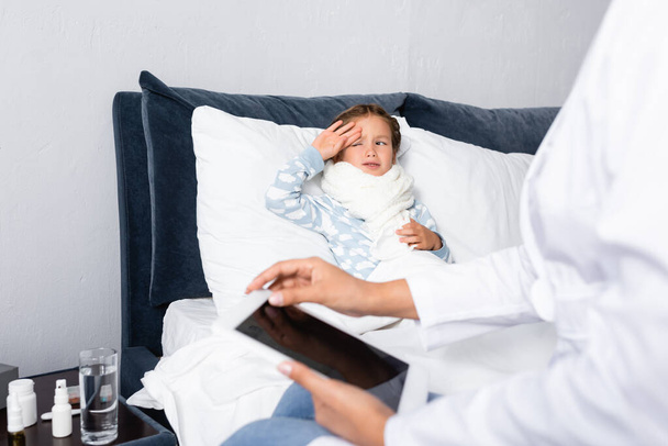 pediatra trzymający tabletkę cyfrową z pustym ekranem w pobliżu chorego dziecka leżącego w łóżku - Zdjęcie, obraz