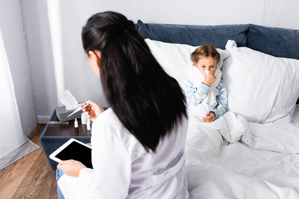 γιατρός κοιτάζοντας θερμόμετρο κοντά άρρωστος κορίτσι που βρίσκεται στο κρεβάτι - Φωτογραφία, εικόνα