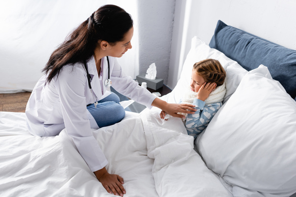 lékař dotýkající se dítěte trpícího bolestí ucha při ležení v posteli - Fotografie, Obrázek