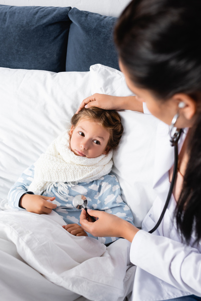 médico tocando cabeça de menina doente, deitado na cama, e examiná-la com estetoscópio - Foto, Imagem