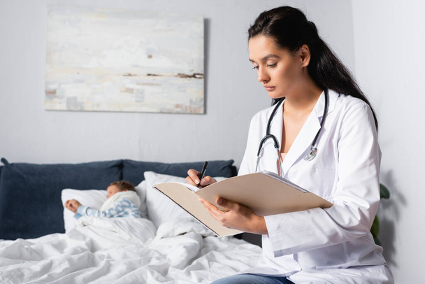 serious doctor writing diagnosis near sick child lying in bed - Valokuva, kuva