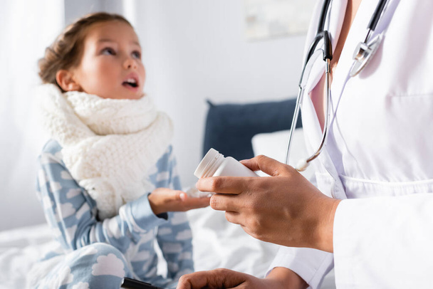 pediatrician giving pills to diseased girl with warm scarf on neck on blurred background - Photo, Image