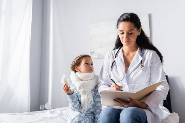 παιδίατρος γράφοντας διάγνωση ενώ άρρωστο κορίτσι κρατώντας χαρτοπετσέτα - Φωτογραφία, εικόνα