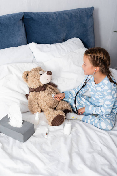 κορίτσι εξέταση αρκουδάκι με στηθοσκόπιο, ενώ παίζει στο κρεβάτι - Φωτογραφία, εικόνα