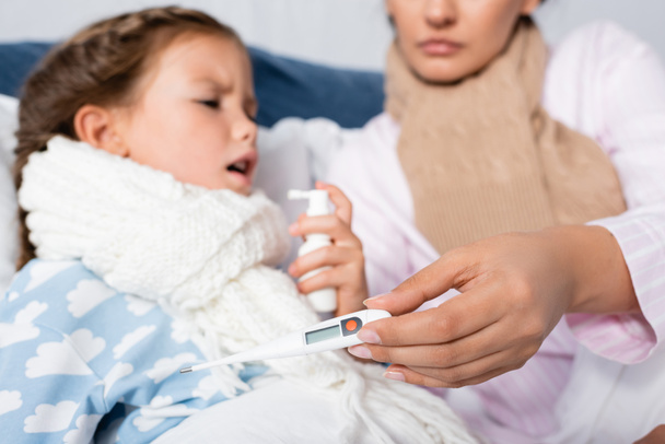 больная женщина держит термометр в то время как дочь использует спрей для горла на размытом фоне - Фото, изображение