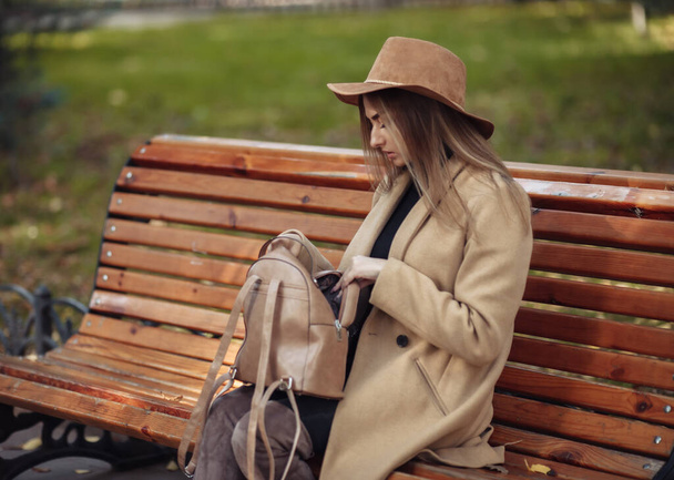 Young attractive woman in autumn clothes sits on bench in city park. Woman dressed in stylish coat and felt hat. Autumn time - Фото, зображення