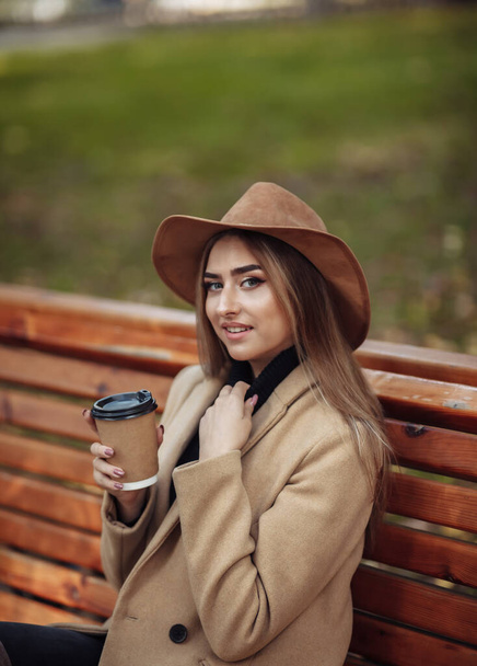 Young attractive woman in autumn clothes sits on bench and drinks coffee in city park. Woman dressed in stylish coat and felt hat. Autumn time - Photo, Image