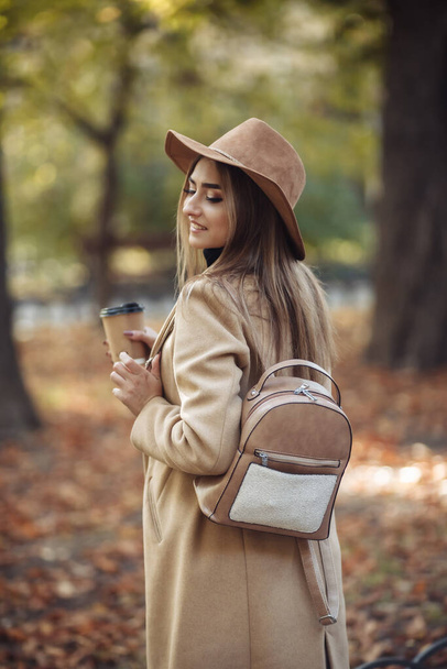 Young attractive woman in autumn clothes and drinks coffee while walking in city park. Woman dressed in stylish coat and felt hat. Autumn time - Fotografie, Obrázek