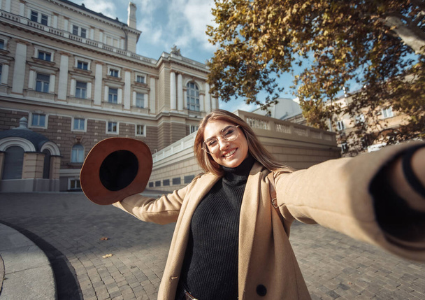 Selfie portret van een jonge stijlvolle toeristische vrouw gekleed in een jas en hoed op een achtergrond van de Europese stedelijke architectuur. Vakantie en toerisme concept - Foto, afbeelding