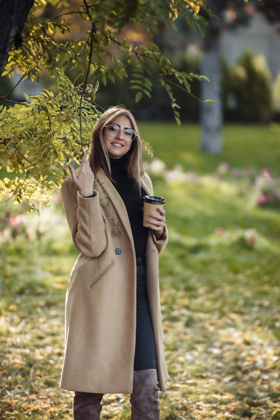 Mujer atractiva joven vestida con un abrigo elegante, cuello alto posando en el parque de otoño y sostiene la taza de café - Foto, imagen