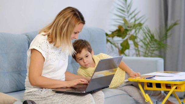 Usmívající se máma pracuje doma se svým dítětem na gauči, zatímco píše e-mail. Mladá žena pracuje z domova, zatímco je v karanténě. Klidná mladá matka nebo chůva sedět na gauči pracuje na notebooku - Fotografie, Obrázek