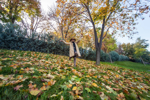 Jeune femme drôle vêtue d'un manteau et chapeau vomit des feuilles tombées dans le parc d'automne. Amusement d'automne - Photo, image