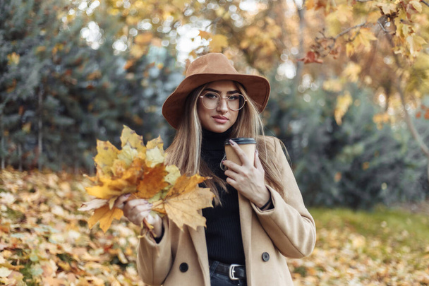 コートや帽子に身を包んだ若い面白い女性は、秋の公園で落ち葉の軍隊を保持しています。秋の楽しみ - 写真・画像