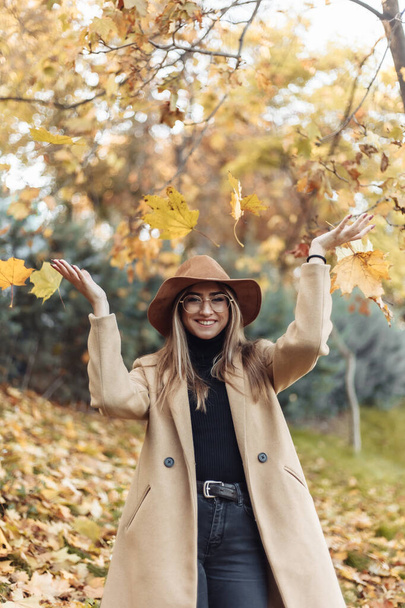 Young funny woman dressed in a coat and hat throws up fallen leaves in the autumn park. Autumn fun - Foto, immagini