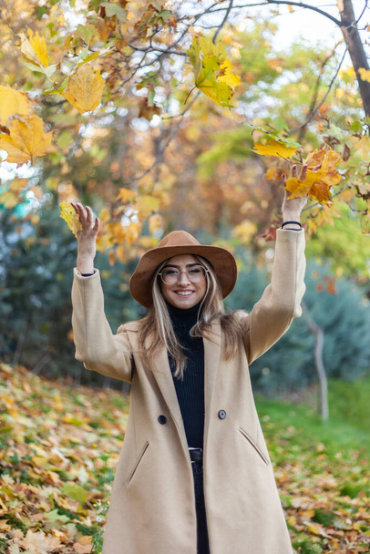 Joven mujer divertida vestida con un abrigo y sombrero arroja hojas caídas en el parque de otoño. Diversión otoñal - Foto, imagen