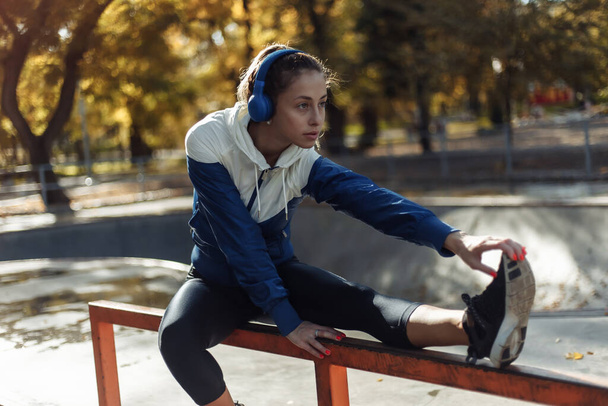 Fit slim woman in sportswear and headphones practicing leg stretching before training in a sports park - Photo, image