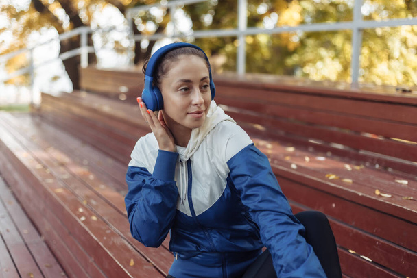 Femme en forme gaie en vêtements de sport assis sur des stands en bois et écoutant de la musique sur des écouteurs. Concept de relaxation d'entraînement - Photo, image