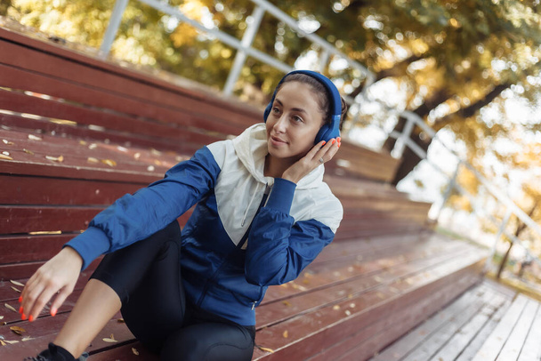 Cheerful fit woman in sportswear sitting on wooden stands and listening to music on headphones. Workout relaxation concept - Photo, Image