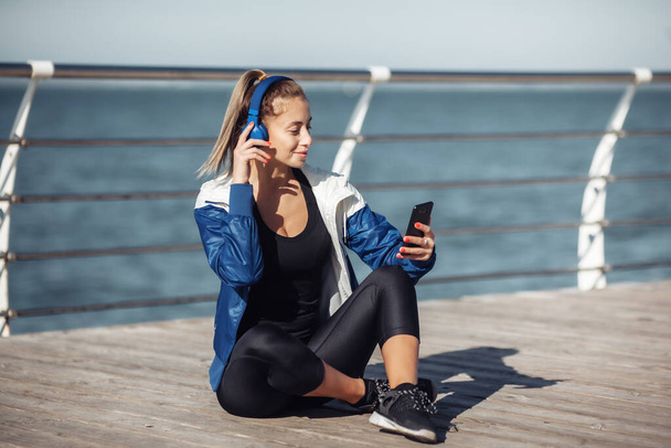 Mujer en forma relajada en ropa deportiva con auriculares elige la música de un teléfono inteligente para el entrenamiento en la playa en un día soleado brillante - Foto, Imagen
