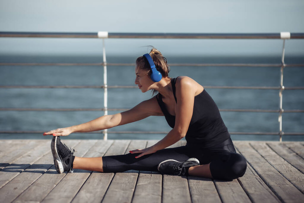 Стройная женщина в спортивной одежде делает растяжку ног и слушает музыку в наушниках на пляже в яркий солнечный день - Фото, изображение