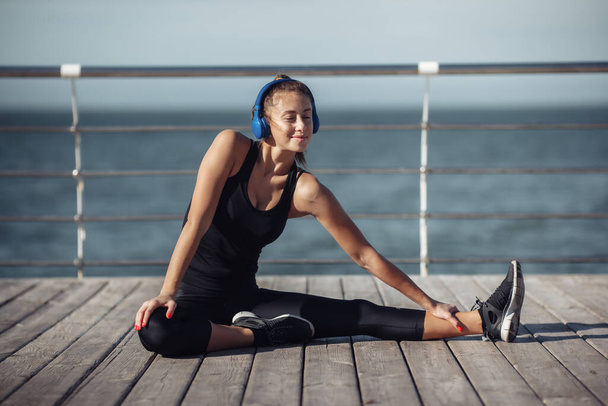 Slim fit γυναίκα στα αθλητικά κάνοντας τέντωμα πόδια και ακούγοντας μουσική με ακουστικά στην παραλία σε φωτεινή ηλιόλουστη μέρα - Φωτογραφία, εικόνα