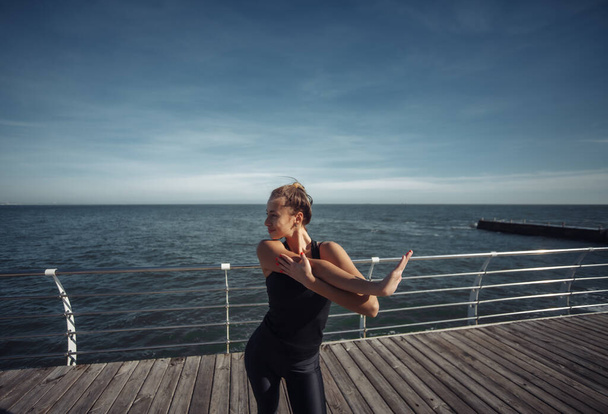 Slim fit vrouw in sportkleding doen stretching hand oefening op het strand op heldere zonnige dag - Foto, afbeelding