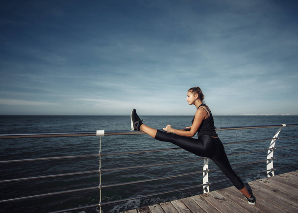 Slim fit kobieta w odzieży sportowej robi ćwiczenia rozciągające nogi na plaży w jasny słoneczny dzień - Zdjęcie, obraz