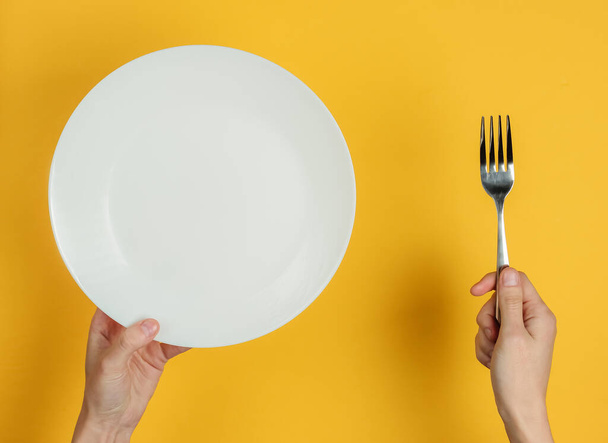 Minimalistický koncept jídla. Ženské ruce držící vidličku a prázdný bílý talíř na žlutém pozadí. Horní pohled. Plocha - Fotografie, Obrázek